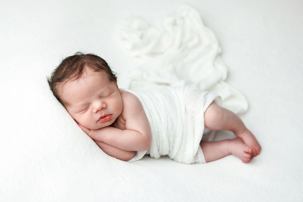 Peaceful newborn baby sleeping wrapped in white cloth on a soft white background.