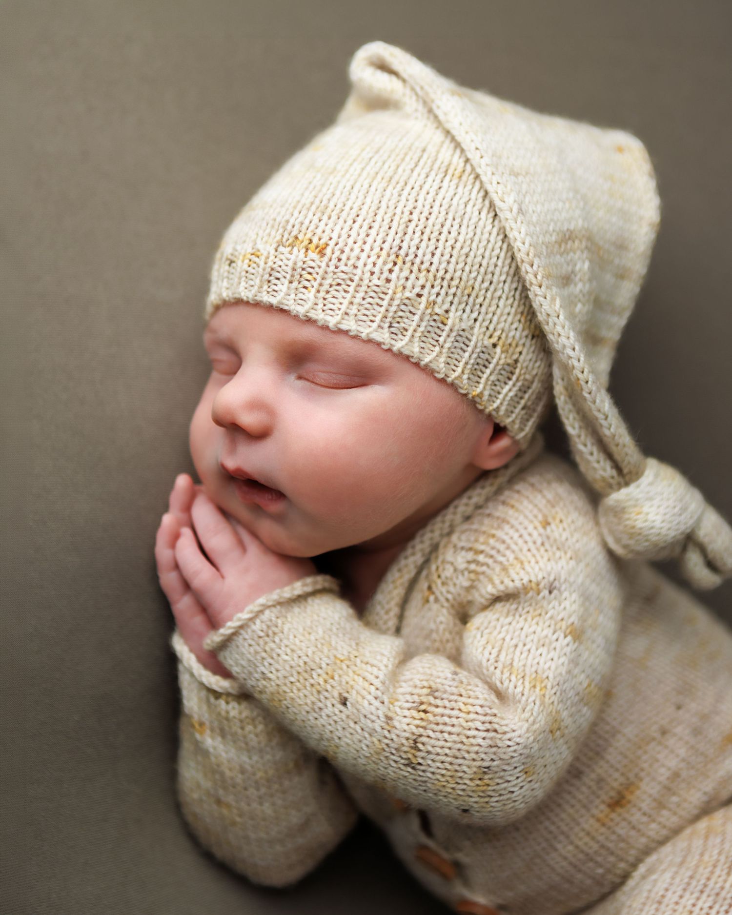Sleeping newborn baby wearing a cream knit romper and matching pointed hat, with tiny hands tucked under chin.