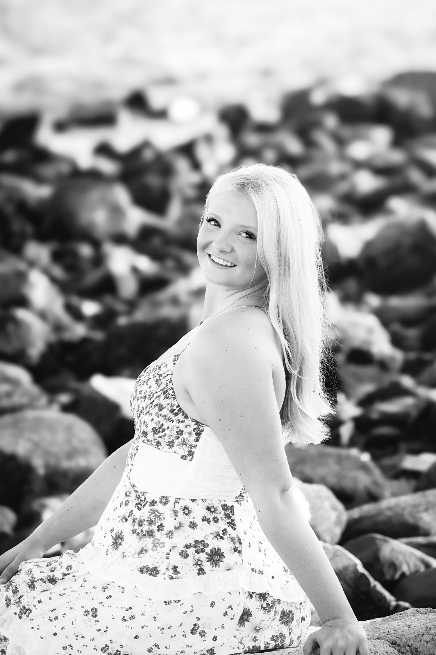 Black and white portrait of someone in a floral dress sitting on rocky terrain and smiling at the camera.