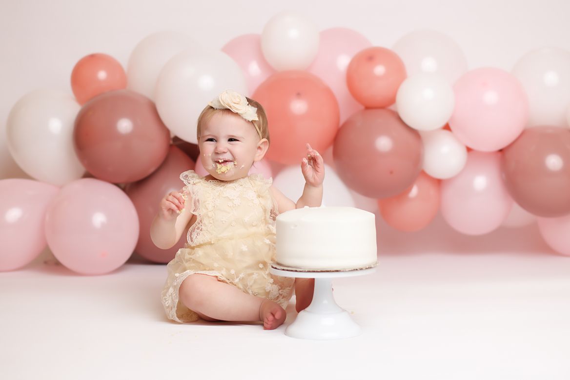 Baby wearing yellow dress sits on white surface with pink and white balloon arch in background.