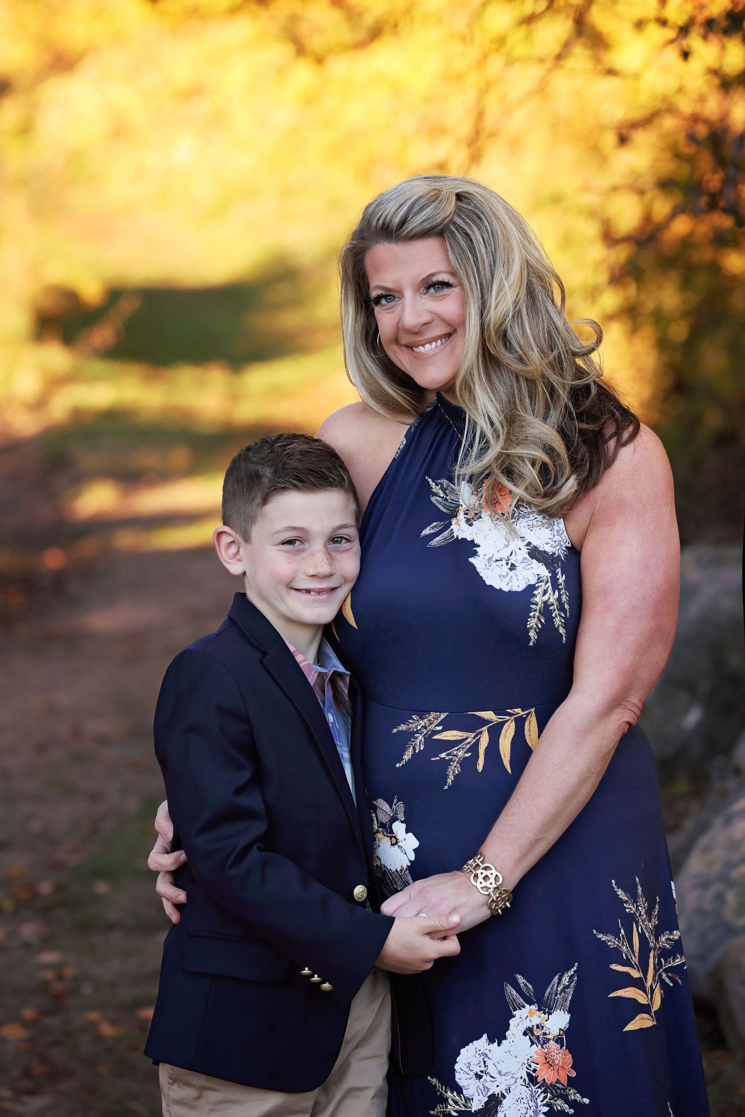 A child in navy blazer embraces someone wearing a floral navy dress during an autumn sunset photoshoot.