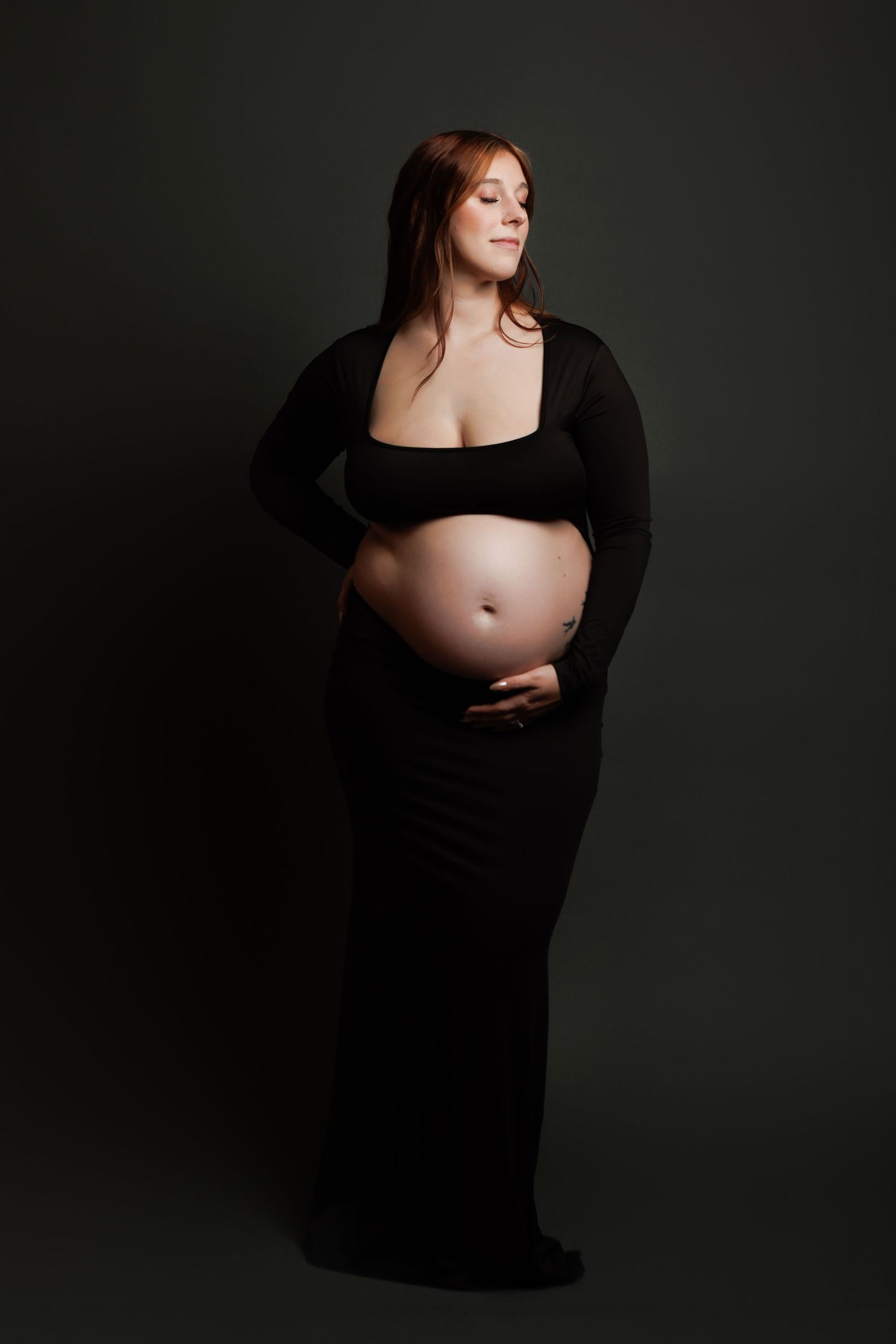 Pregnant woman in black crop top and maxi skirt cradling her bare baby bump against a dark studio background.