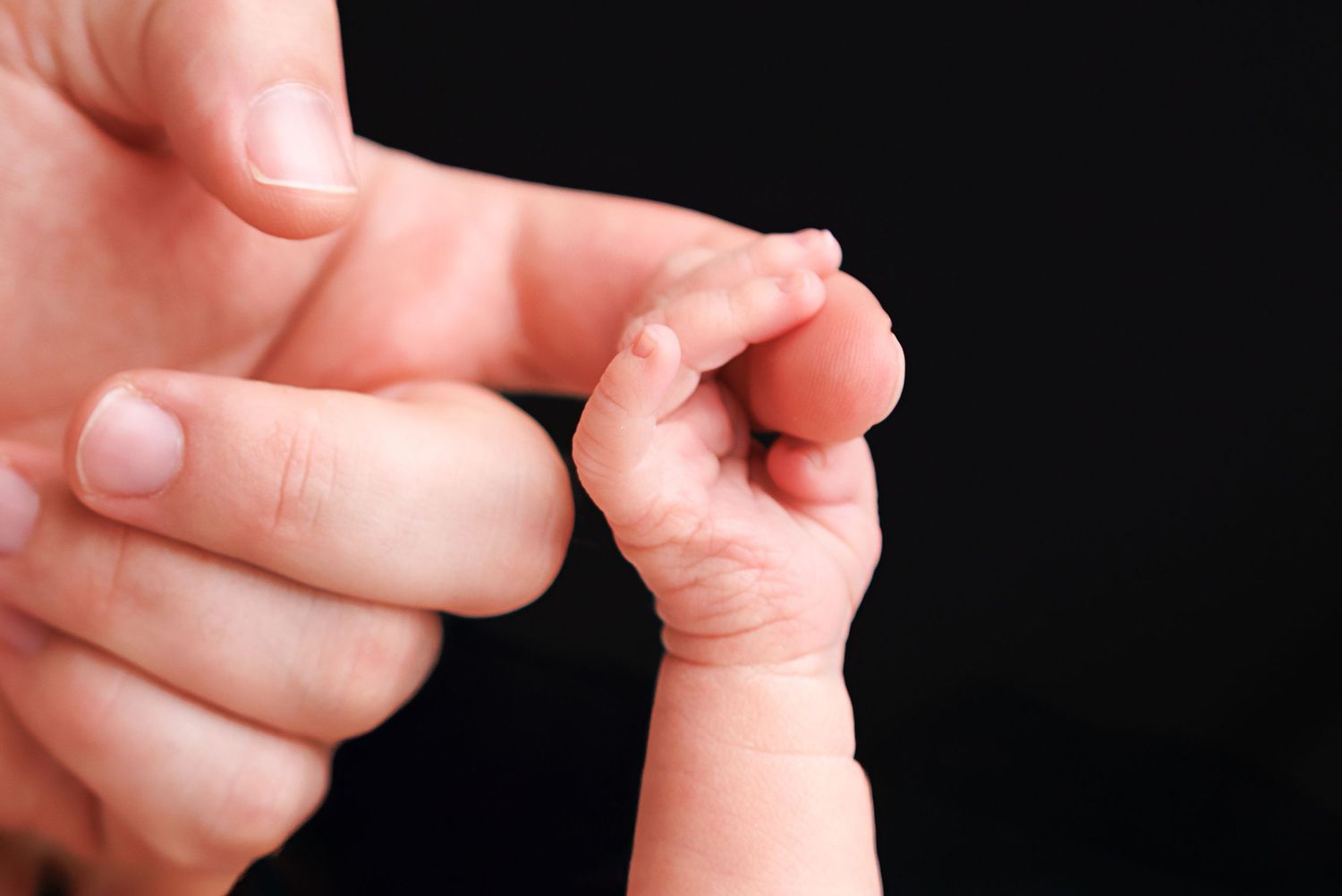 Newborn baby hand grasping adult finger, close-up on black background, symbolizing new life and parental bond.
