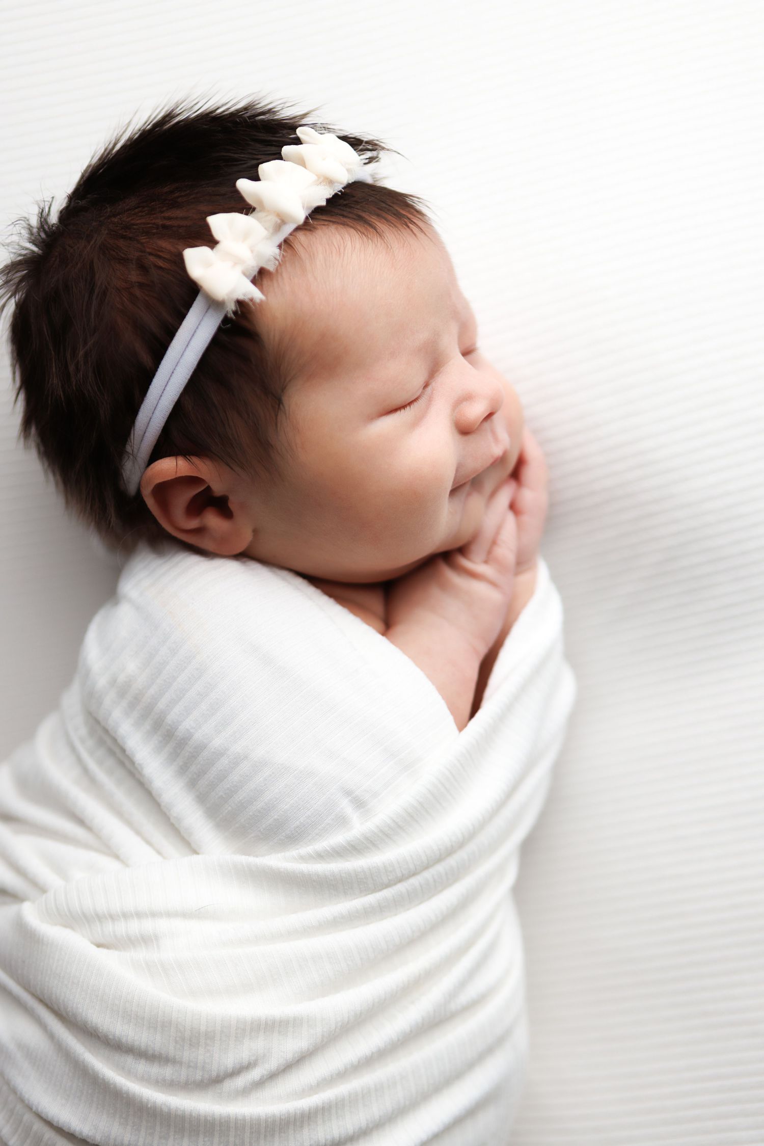 Sleeping newborn baby wrapped in soft white blanket with small bow headband resting peacefully.