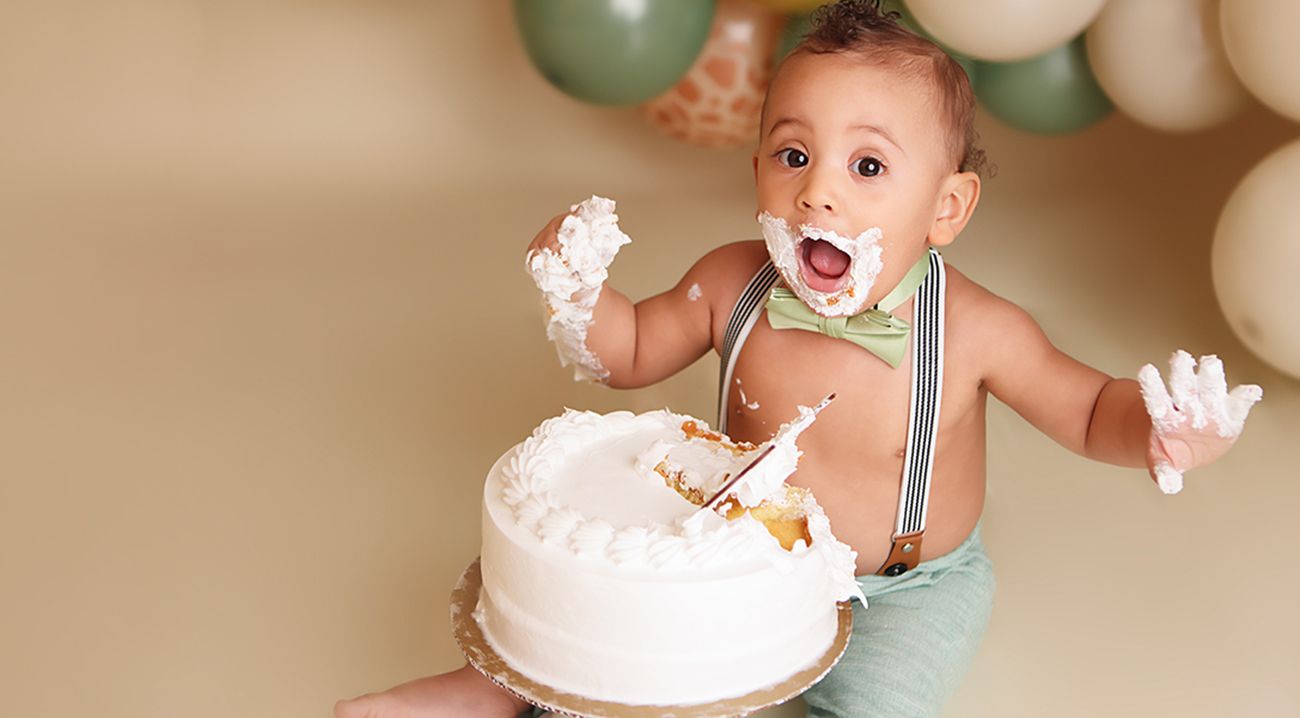 A baby in a light blue outfit smashes and plays with a white birthday cake during a first birthday celebration.