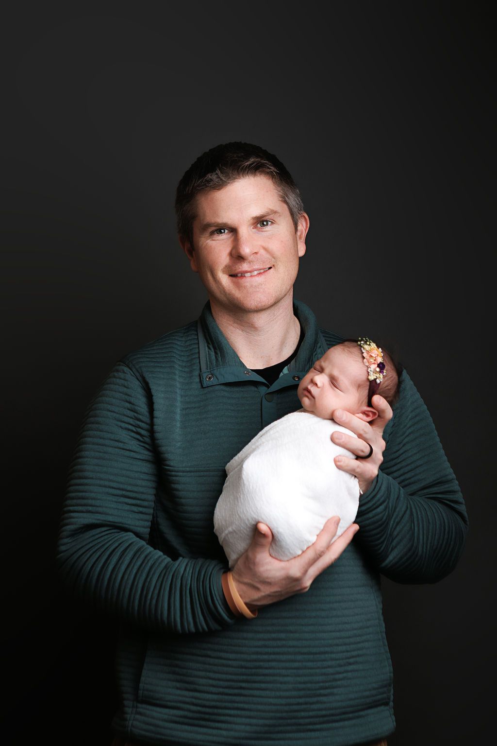 Smiling father in green shirt proudly holds swaddled newborn baby girl with floral headband against dark background.