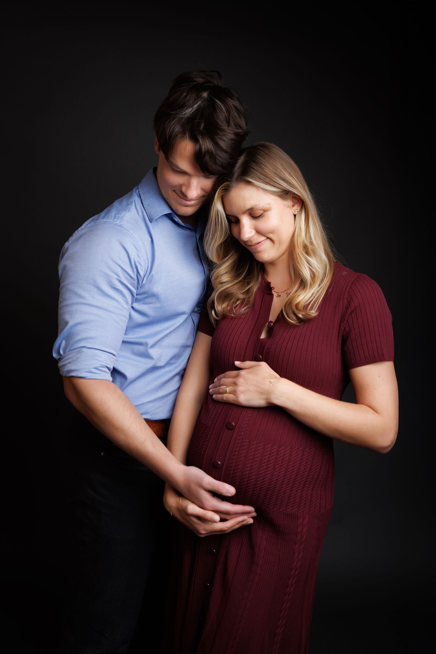 A couple tenderly cradles a pregnant belly during a studio maternity portrait session against a dark background.