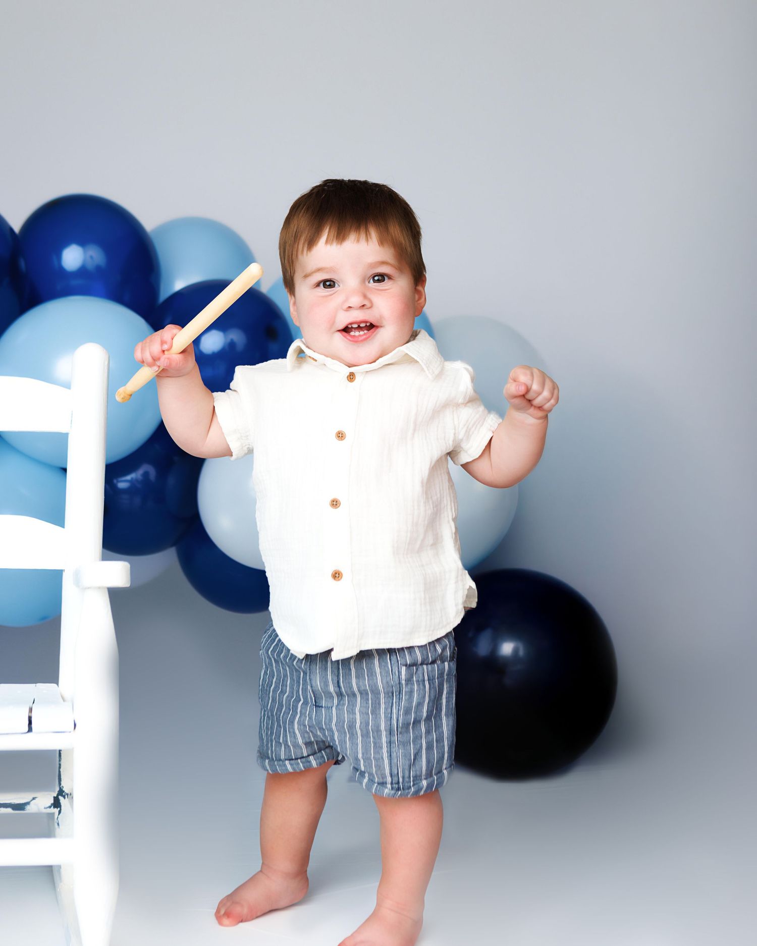 Standing one-year-old boy beside a white chair during first birthday photography in Salem NH