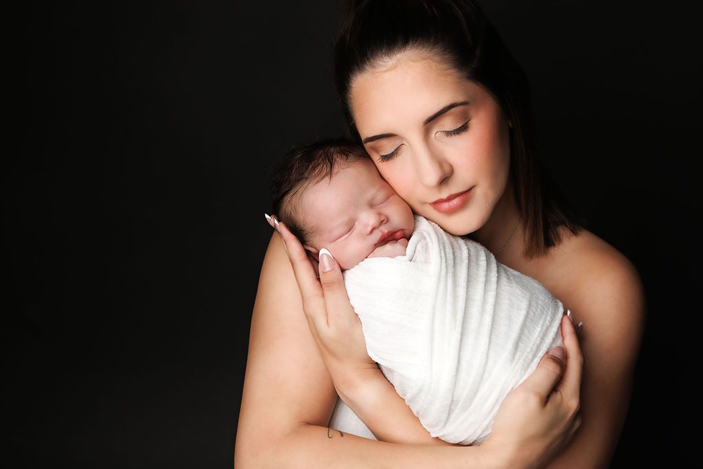 A mother tenderly holds her sleeping newborn wrapped in white against a dark background.