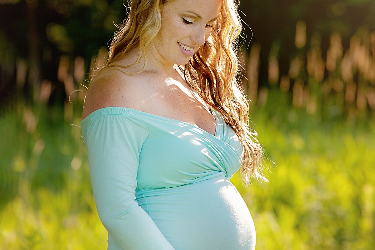 A expectant mother in a light blue dress poses outdoors in a sunny field with tall grass and natural lighting.