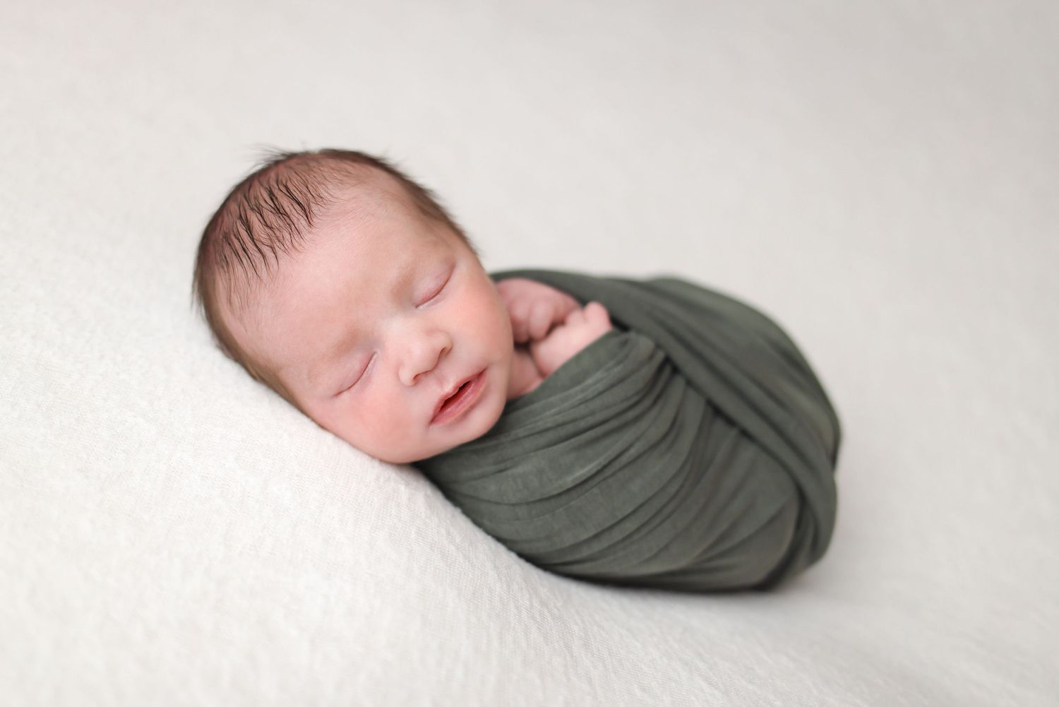 A sleeping newborn baby wrapped in an olive green swaddle blanket against a white background.