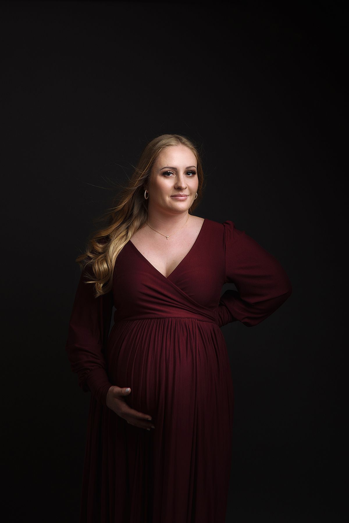 A figure in a burgundy long-sleeve maternity dress poses against a dark background in a series of elegant portraits.