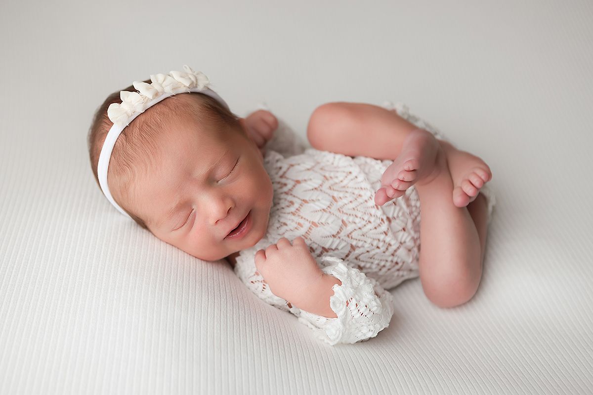 Sleeping newborn baby wearing white lace romper lies peacefully on soft white blanket.