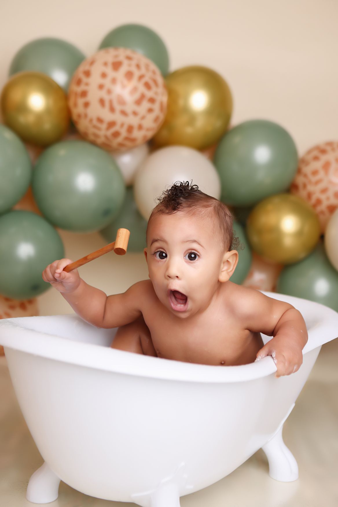Baby splashing in white bathtub with mint green and gold balloons in background during photo session.