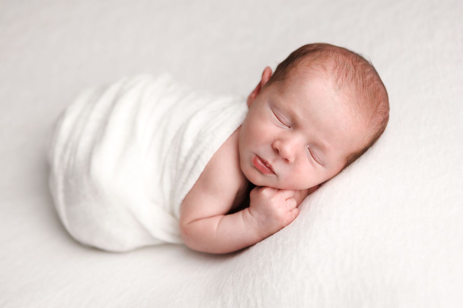 Newborn baby sleeping peacefully, swaddled in a soft white wrap, resting on a white blanket.