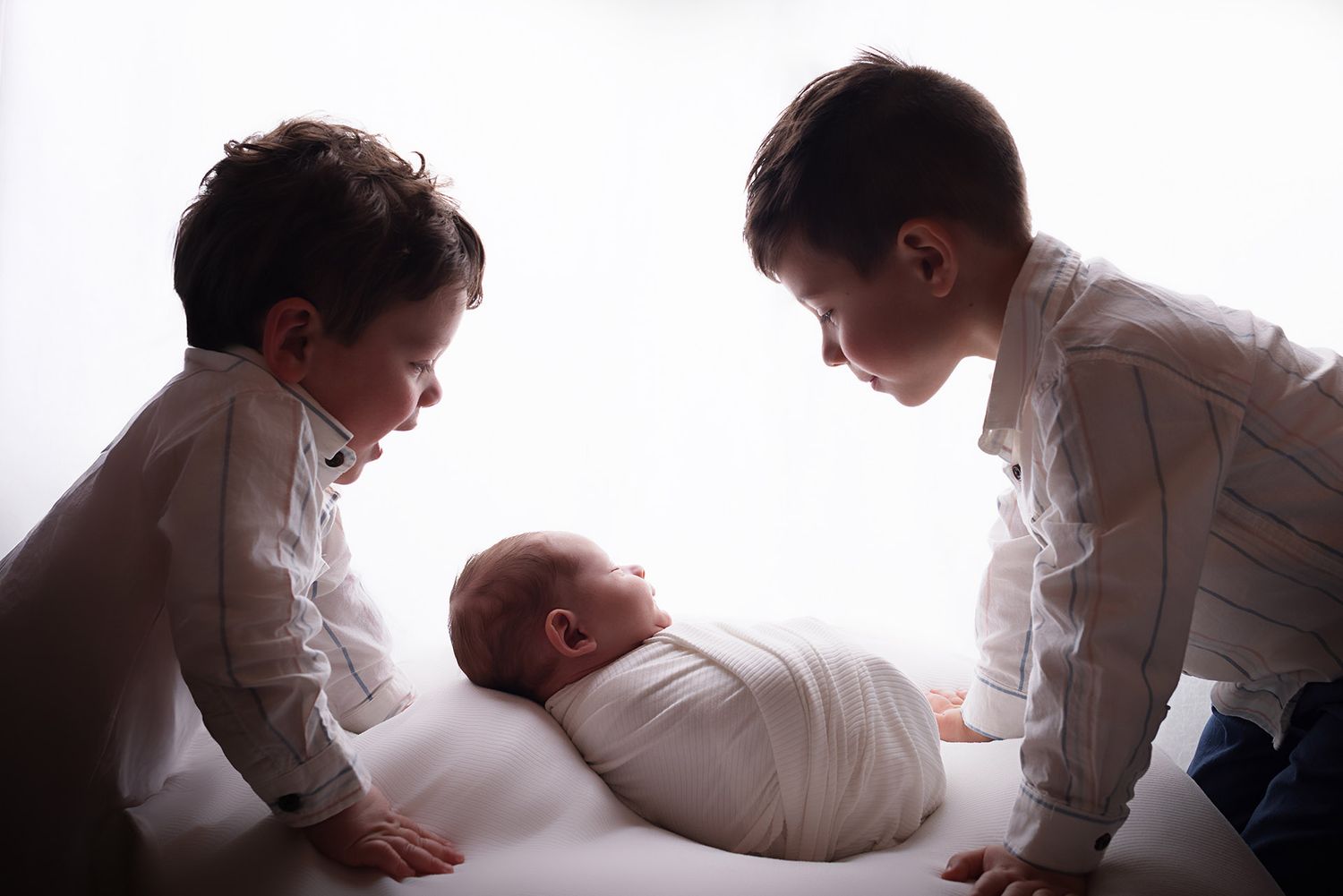Two young boys in white shirts lean over a sleeping newborn baby wrapped in white on a soft surface.