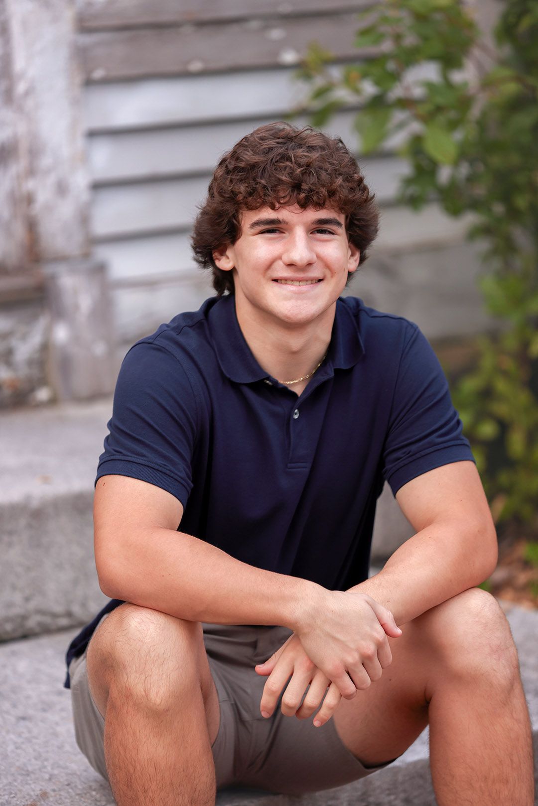 Smiling teenage boy with curly hair sitting on steps outdoors wearing a navy polo shirt and khaki shorts.