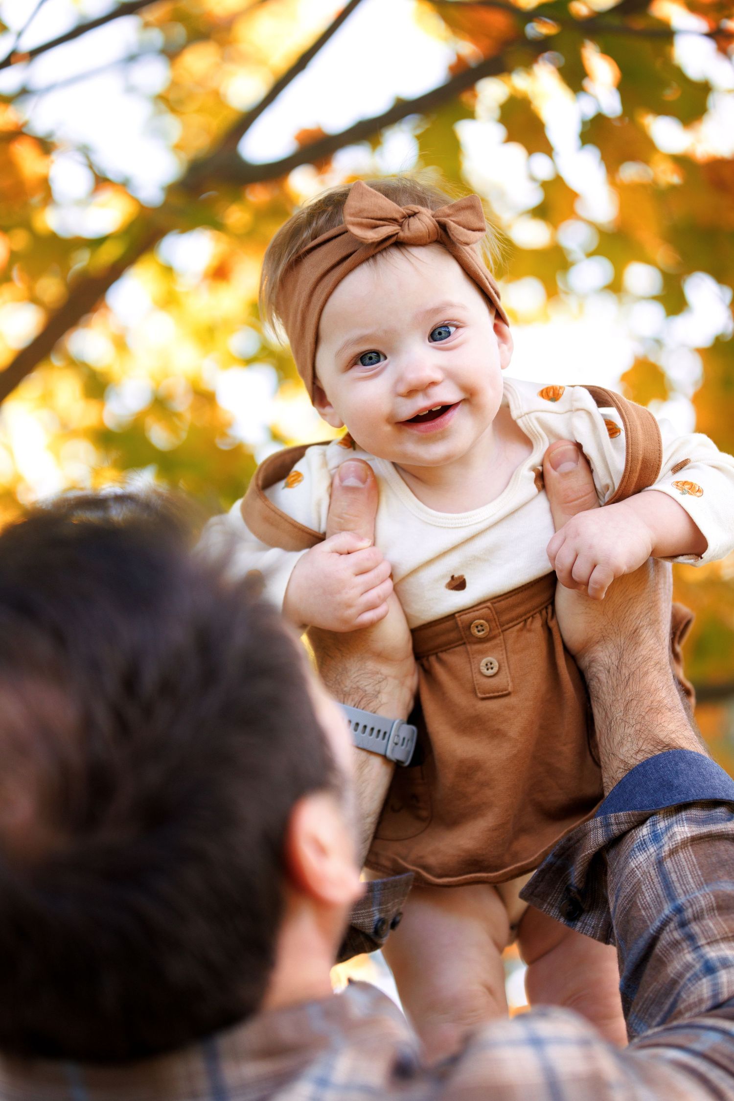 A joyful baby wearing a brown outfit is lifted up against a backdrop of golden autumn foliage.
