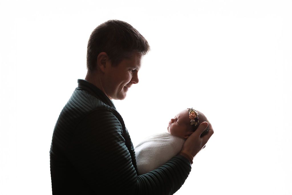 Father holds swaddled newborn baby in silhouette against bright white background.