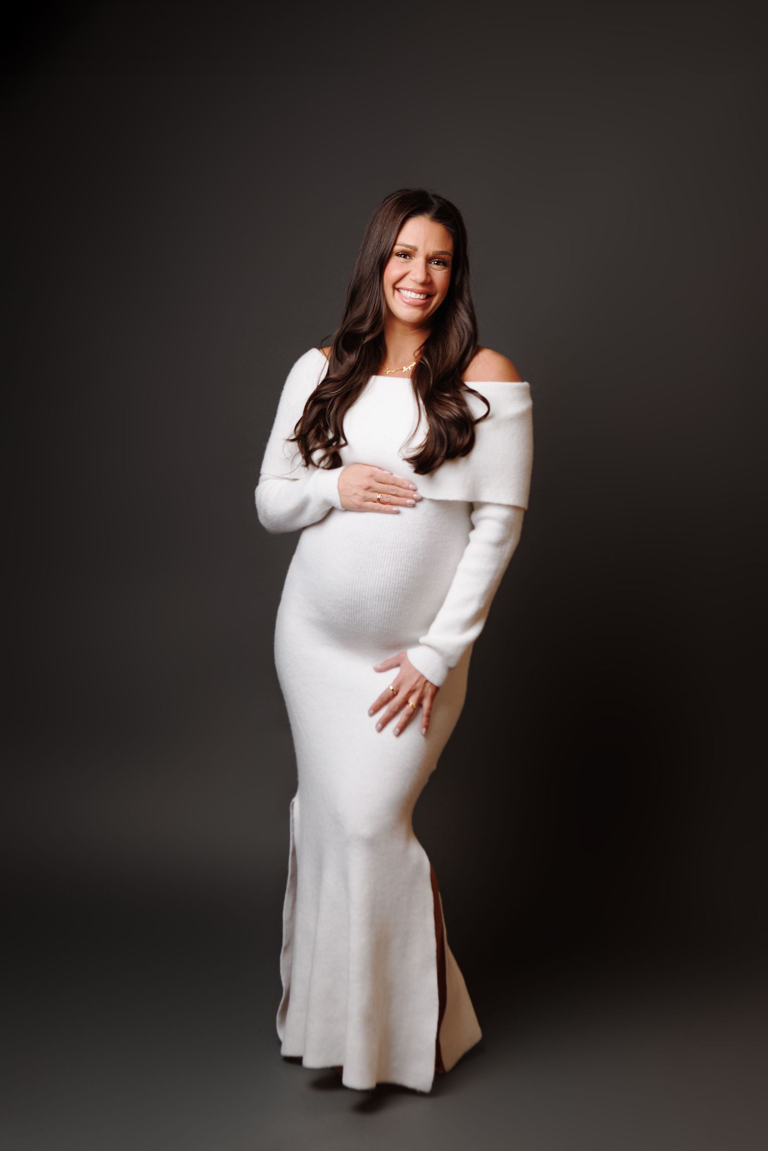Smiling pregnant woman in white off-shoulder knit gown posing against dark background.