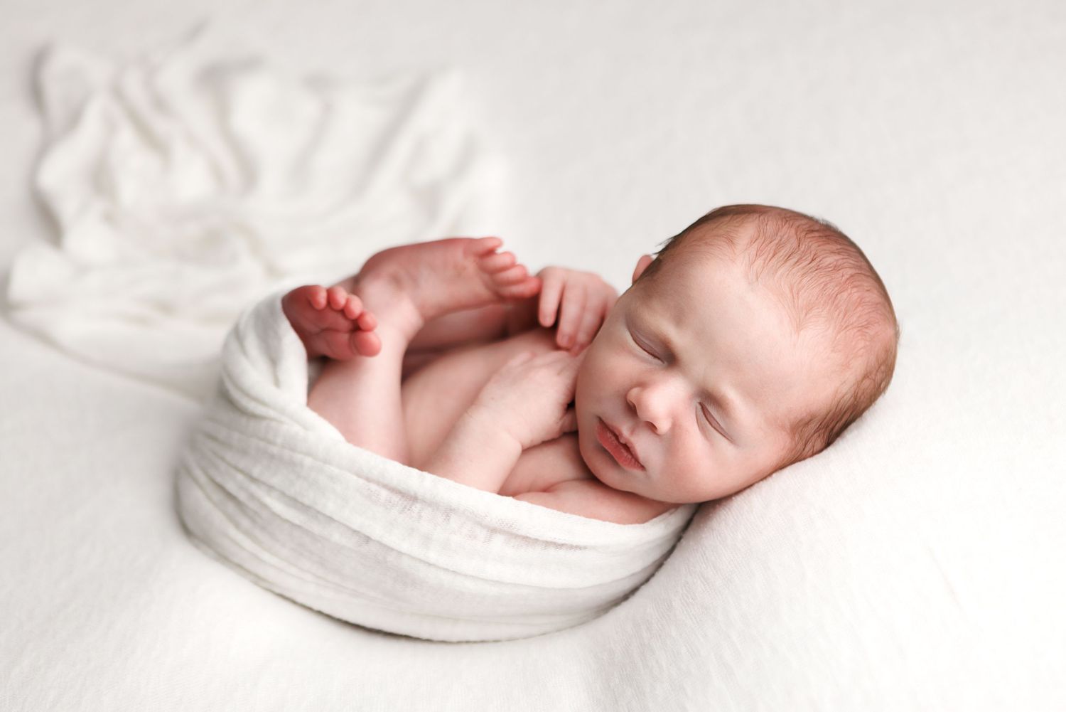 Newborn baby sleeping peacefully wrapped in white cloth, curled up in a cozy pose on a soft white blanket.