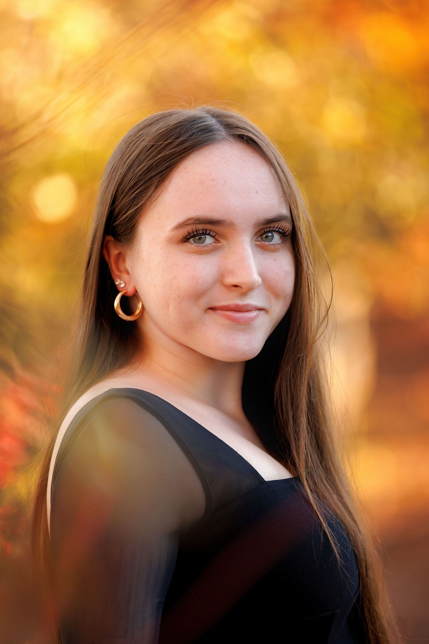 Young woman with long brown hair and green eyes smiling outdoors in autumn, wearing gold hoop earrings and black top.