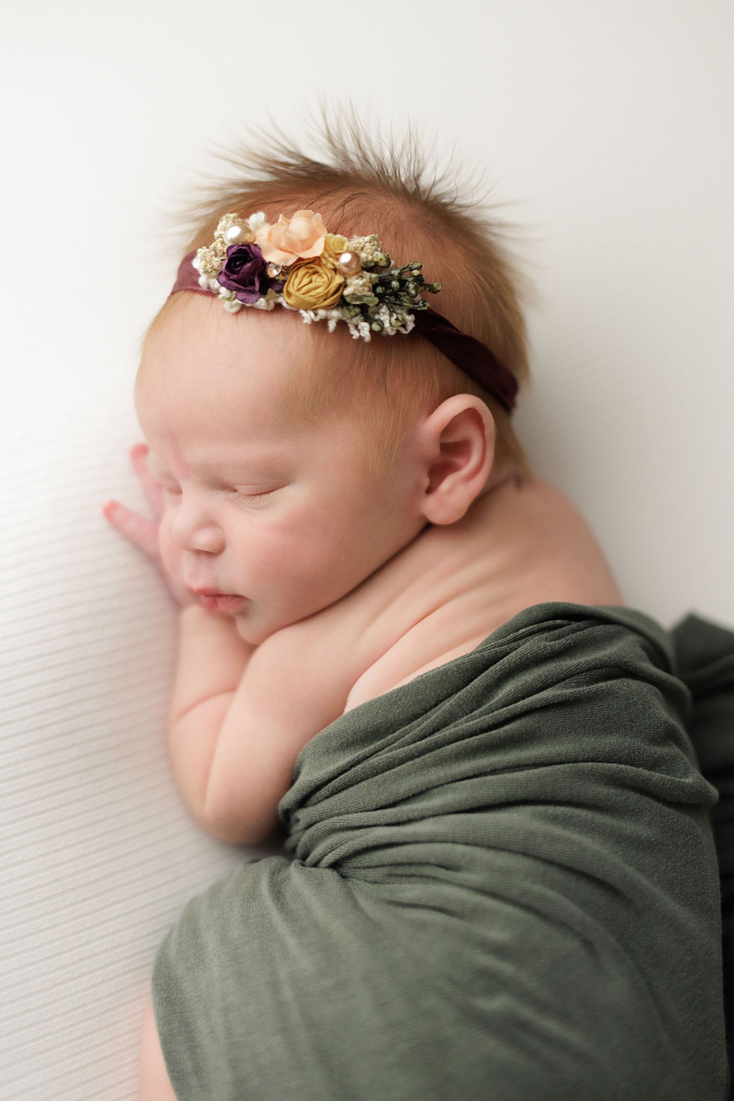 A sleeping newborn baby wrapped in a green swaddle blanket with delicate floral headpiece against white background.