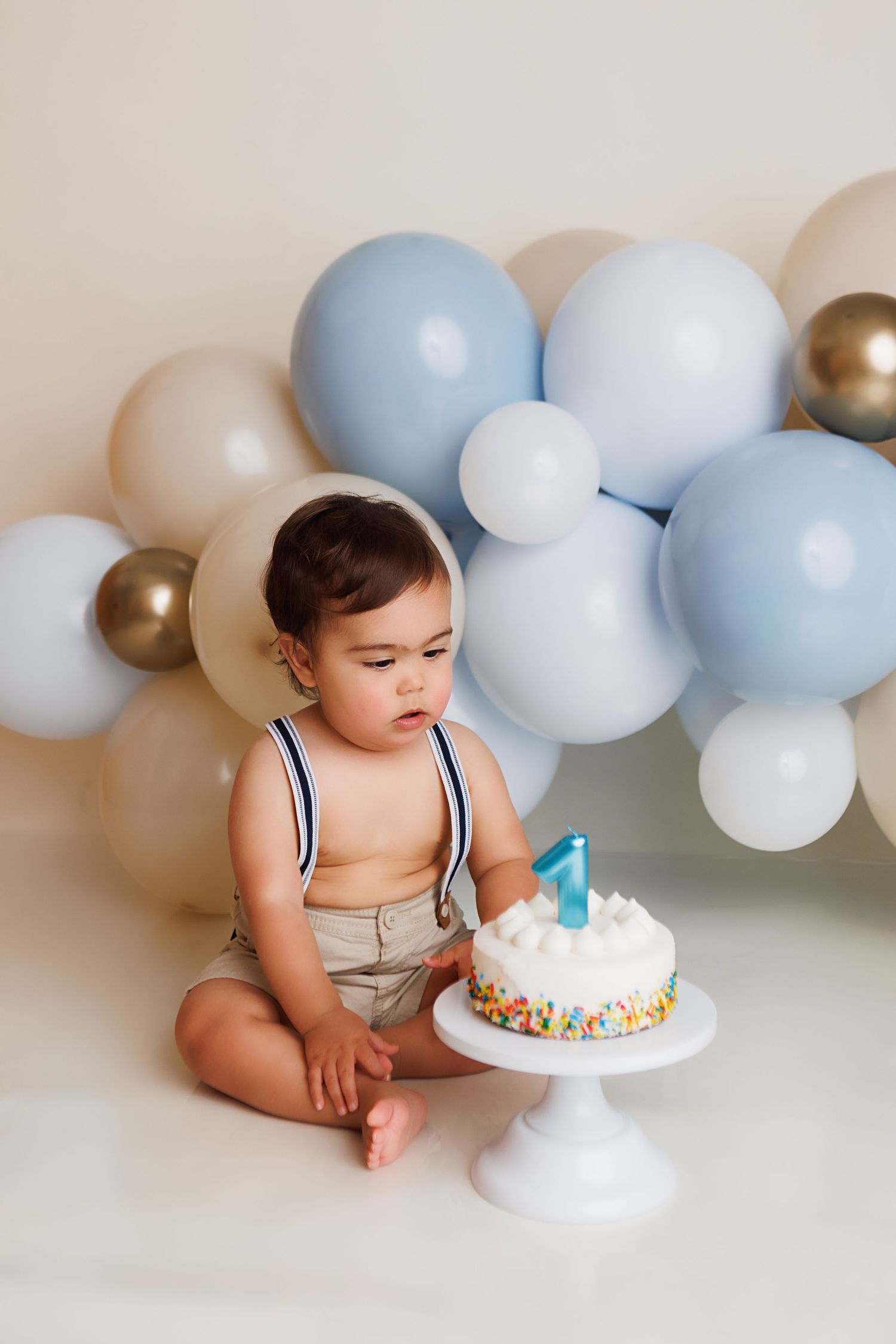 Baby in neutral colored outfit sits beside blue and white balloon garland with birthday cake for cake smash photo session.