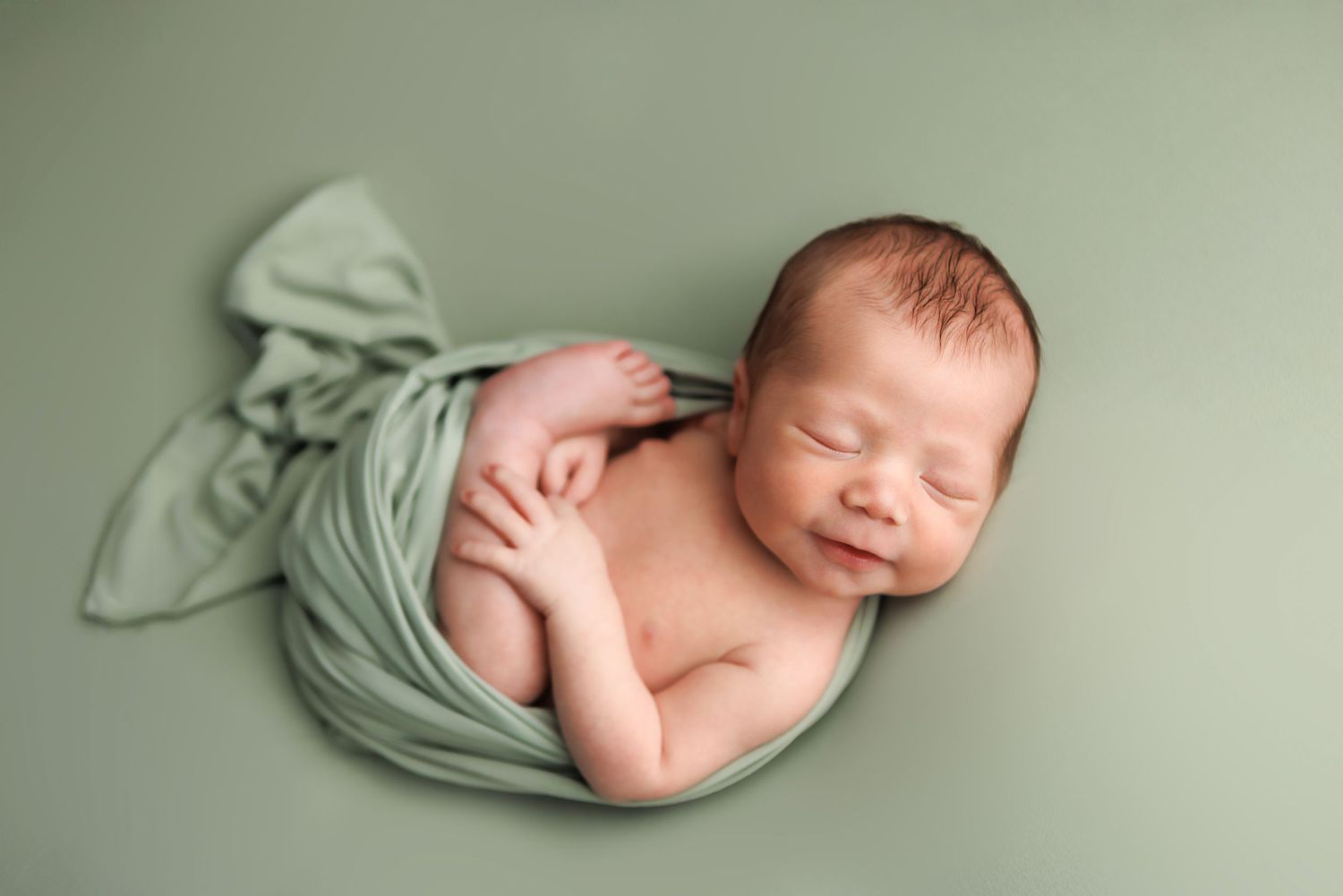 Newborn baby sleeping peacefully on soft sage green blanket in professional studio portrait setting.