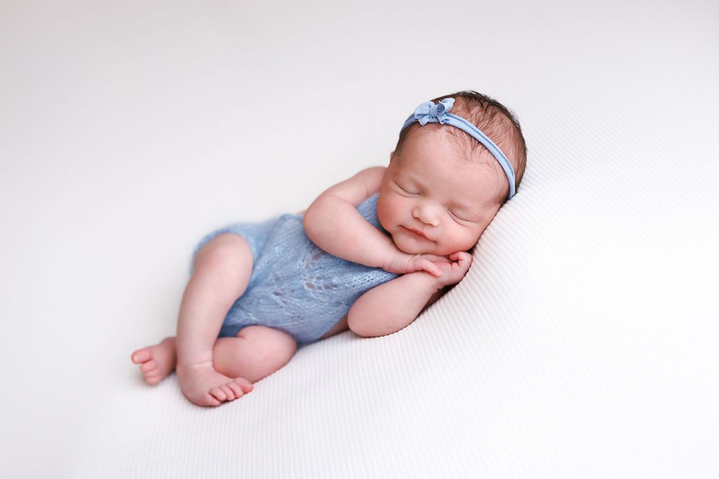 Sleeping newborn baby in blue romper and floral headband posed on white background.