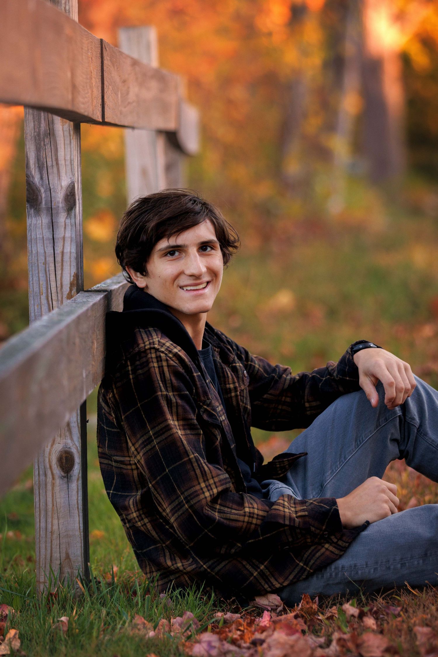 Relaxed autumn portrait session with Senior lounging on wooden fence surrounded by fall leaves.