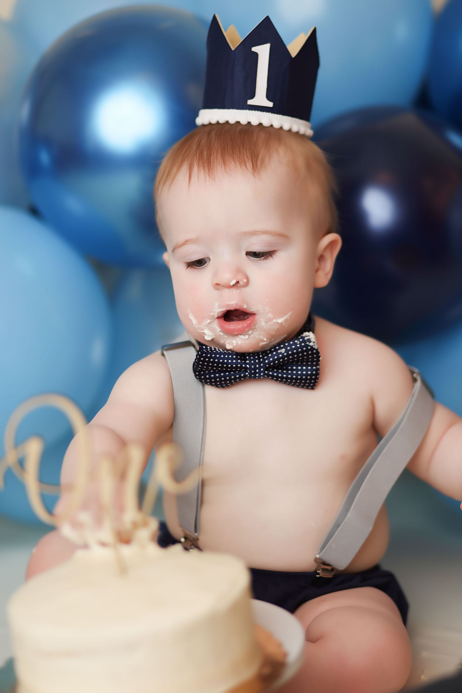 Baby wearing crown, bow tie and suspenders celebrates first birthday with cake against blue balloon backdrop.