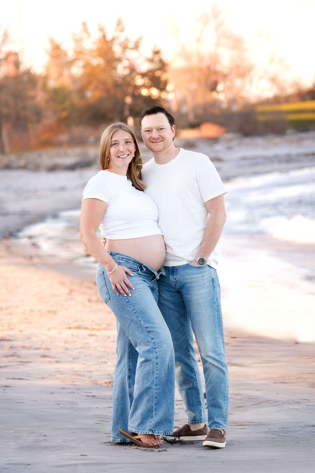 Smiling pregnant couple posing on sandy beach at golden hour in matching white tops and jeans.