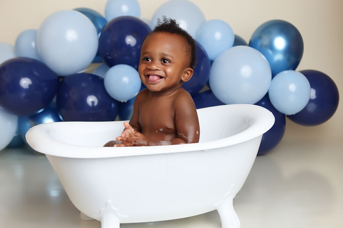 Baby sits in vintage bathtub surrounded by blue balloons during milestone photo session.