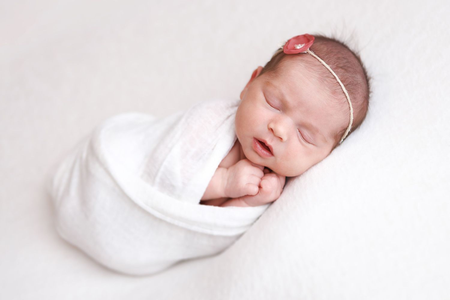Newborn baby girl swaddled in white wrap with pink floral headband, sleeping peacefully on fluffy white background.