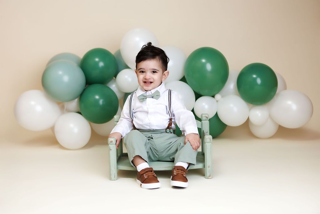 Baby boy in suspenders and bow tie smiles on a small chair surrounded by green and white balloons.