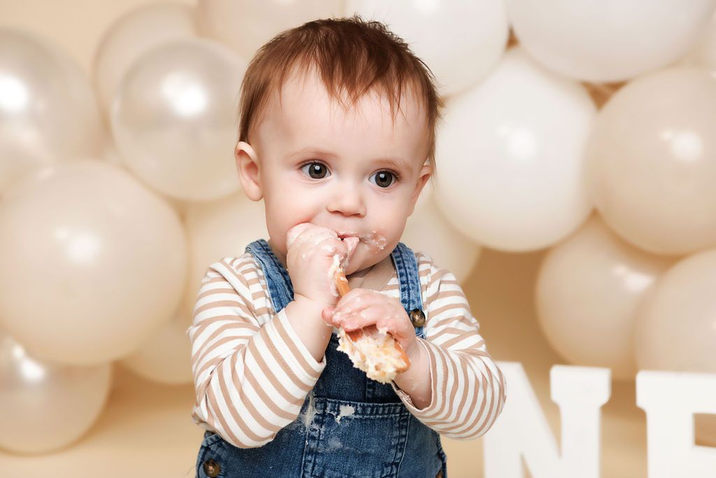 Cute baby in denim overalls eating cake in front of white balloons at first birthday.