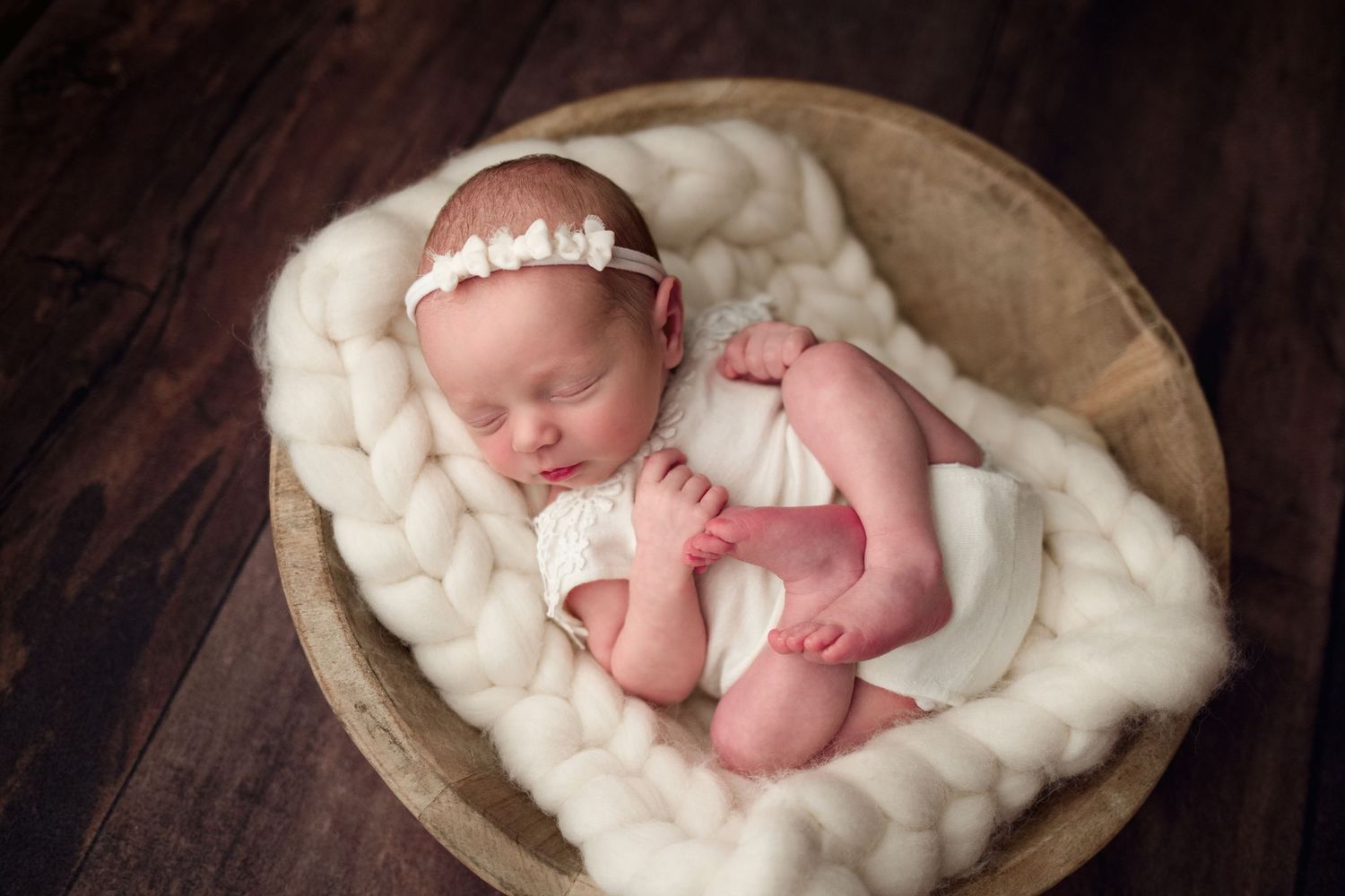 A sleeping newborn baby nestled in soft white blankets inside a rustic wooden bowl against a dark background.