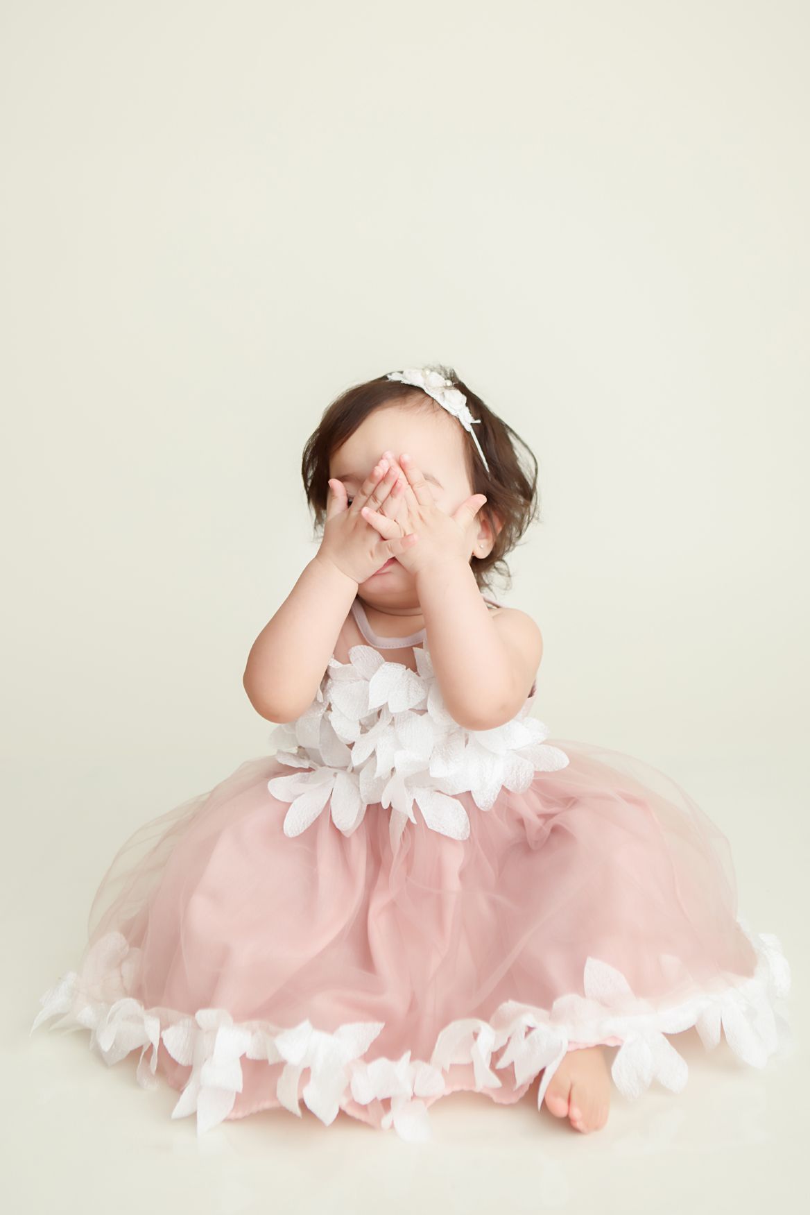 A baby in a pink tulle dress with white flowers plays peek-a-boo in a series of sweet poses. Boston Cake Smash Session.