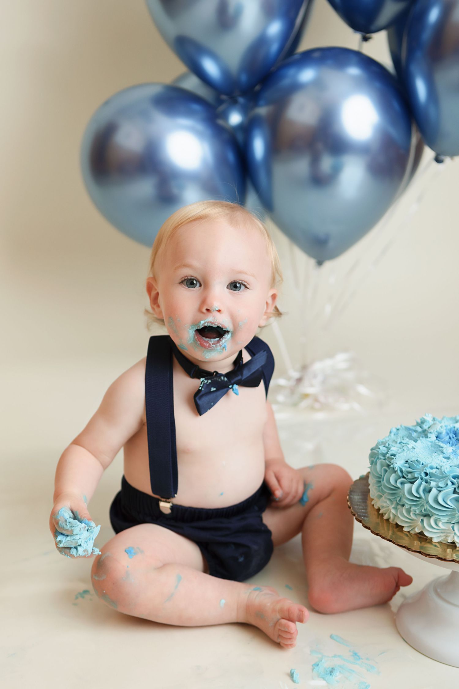 A cake smash photo session with blue balloons and a frosted cake features a baby in navy suspenders and bow tie.