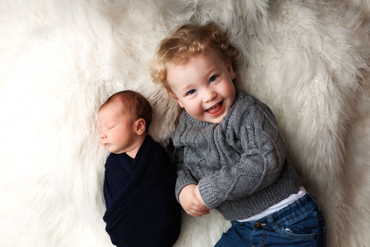 Big Brother and newborn brother laying on white fur backdrop posing for a portrait.