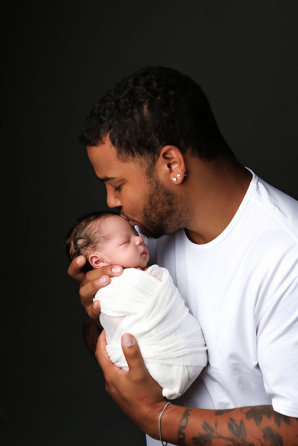 A father tenderly kisses his swaddled newborn baby while holding them gently against a dark background.
