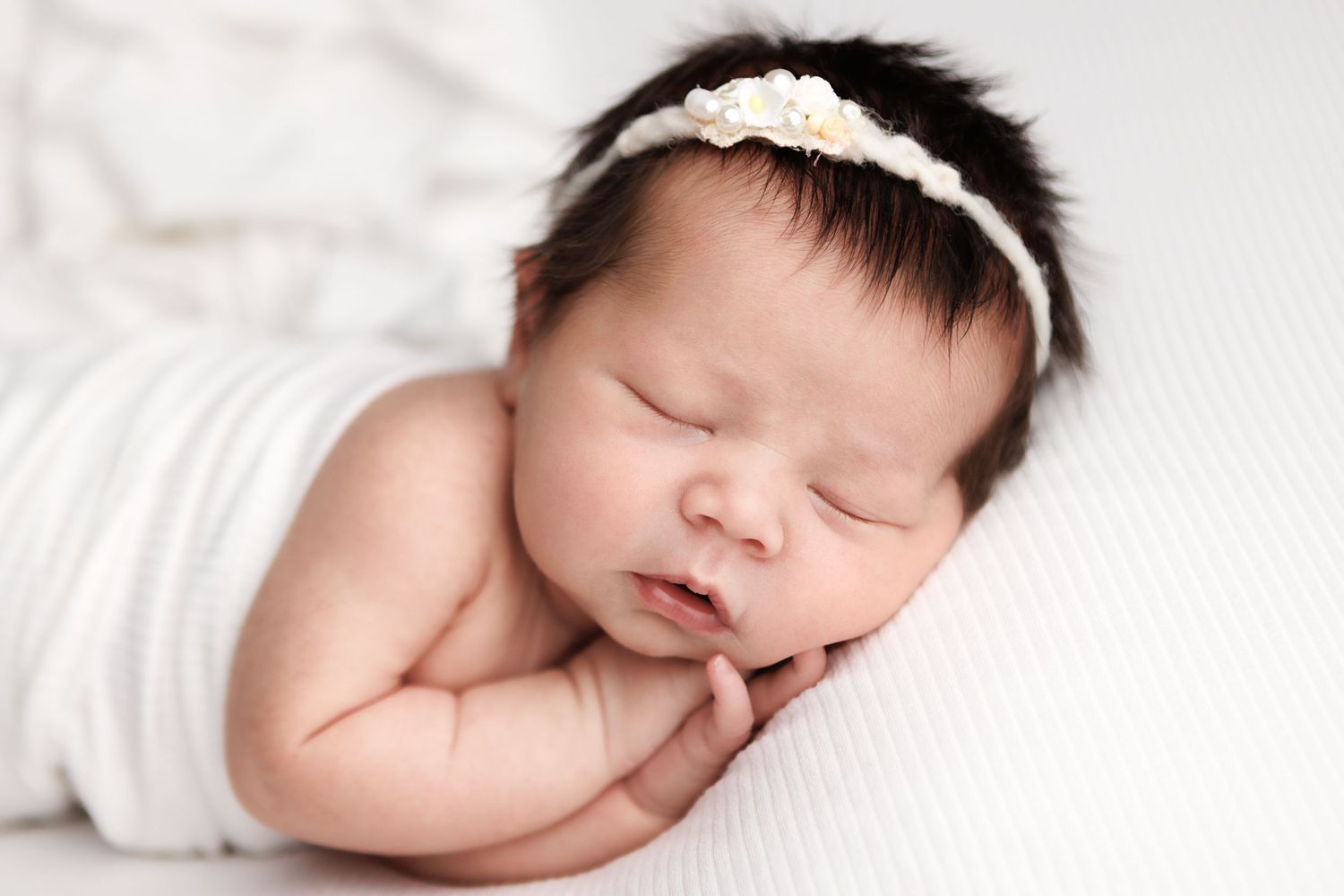 A sleeping newborn baby in white swaddle blanket lies peacefully on white bedding.