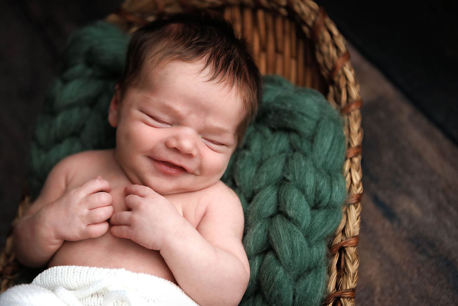 Smiling newborn baby resting in a wicker basket with green chunky knit blanket during professional newborn photography session.