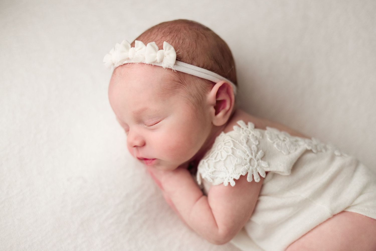 A sleeping newborn baby wearing a delicate white lace headband lies peacefully on a soft cream-colored backdrop.