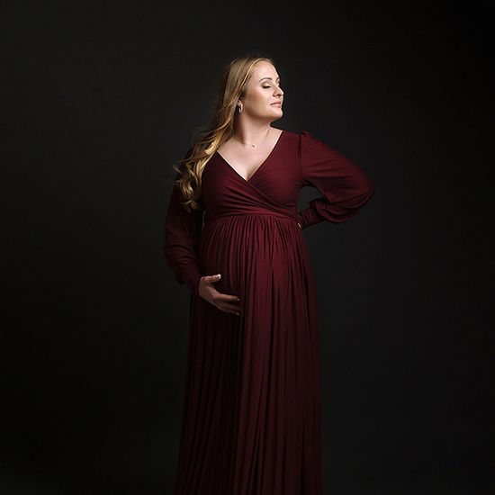 A figure in a burgundy long-sleeved maternity gown poses against a dark backdrop in a series of elegant portraits.