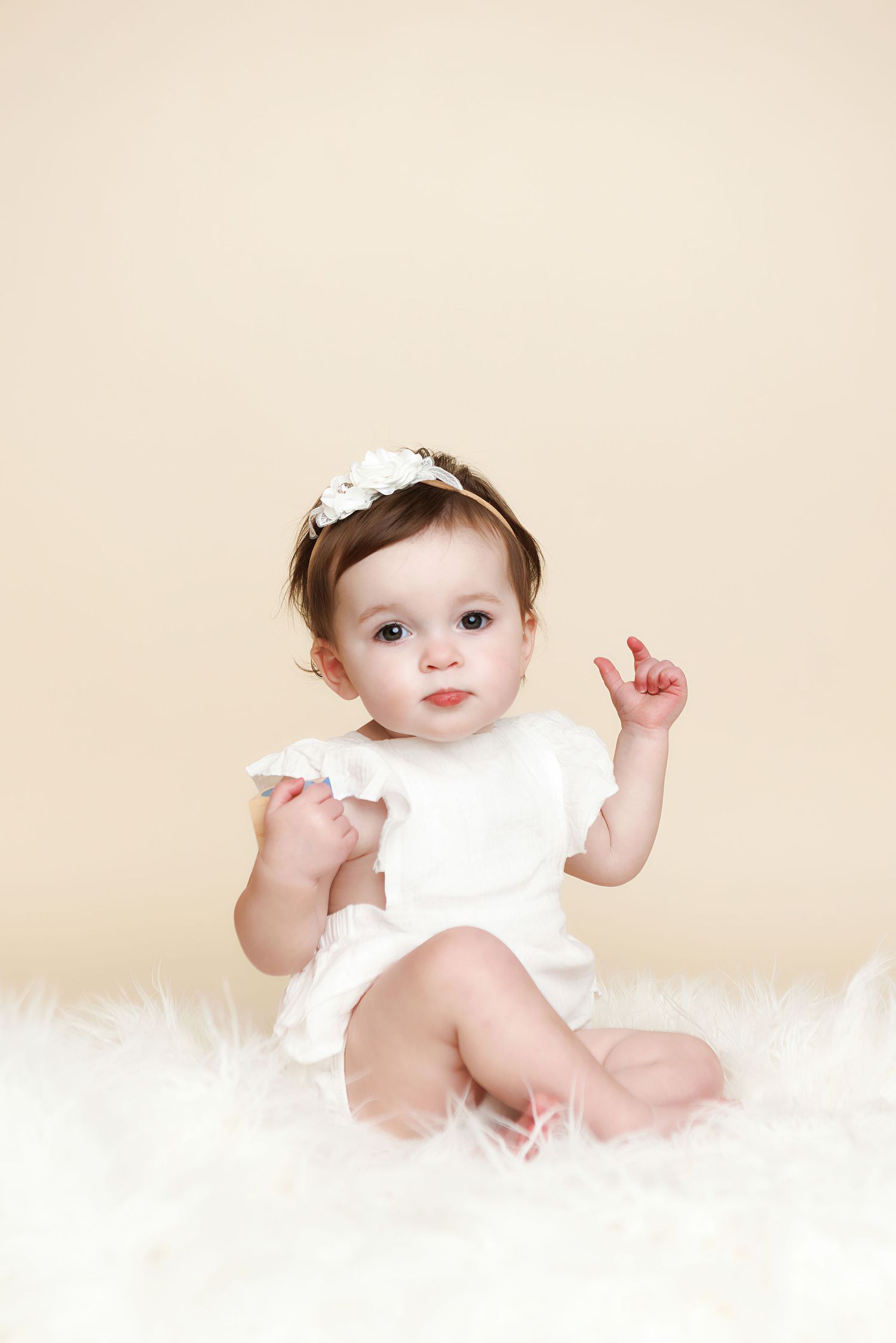 Adorable baby girl in white onesie and floral headband sitting on fluffy white fur rug against beige background.