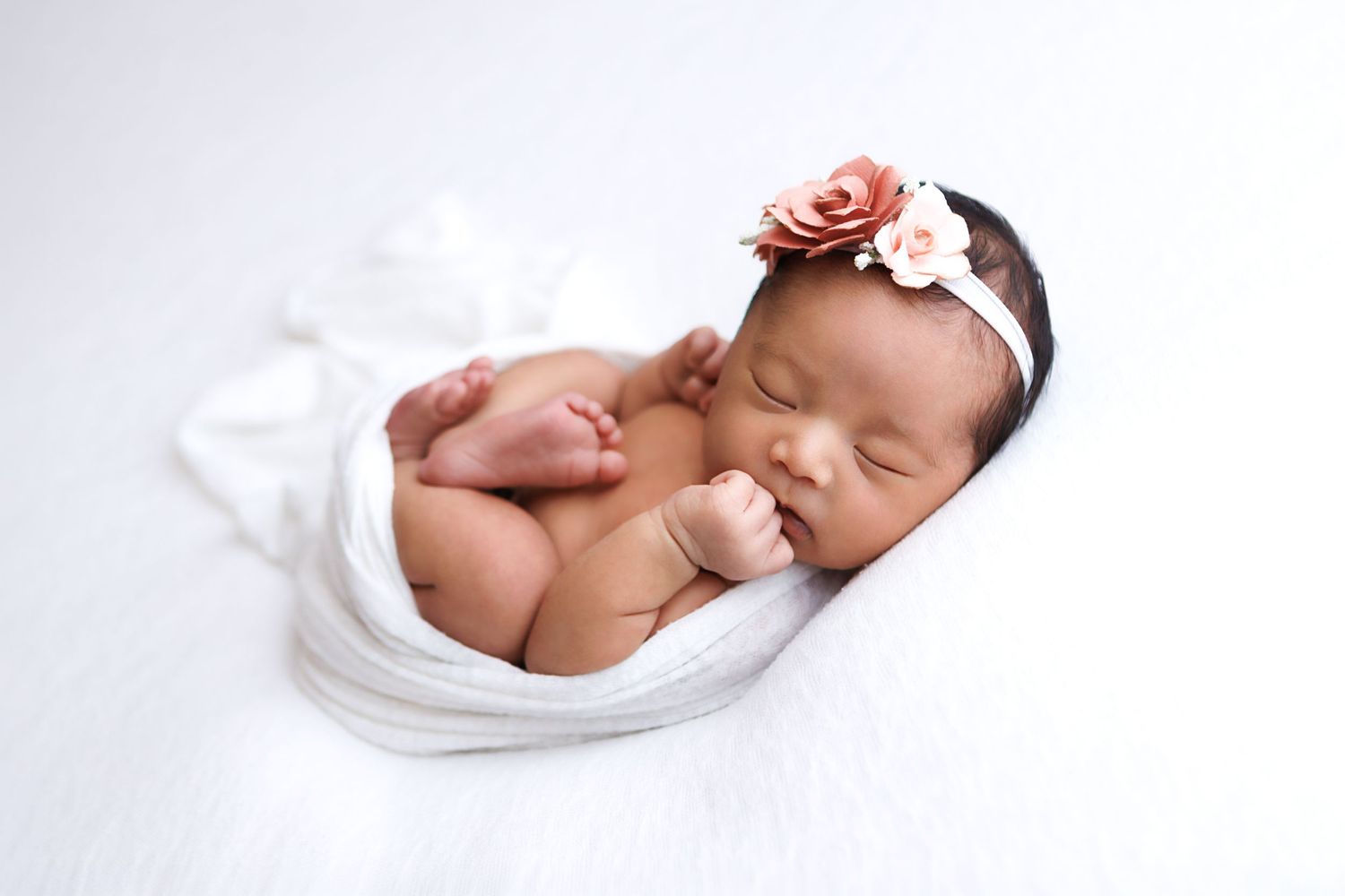 Sleeping newborn baby girl wrapped in white with a pink floral headband on a soft white background.