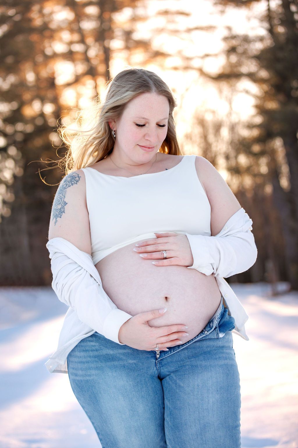 Pregnant woman in white top and jeans cradling baby bump outdoors in snowy winter forest at golden hour.