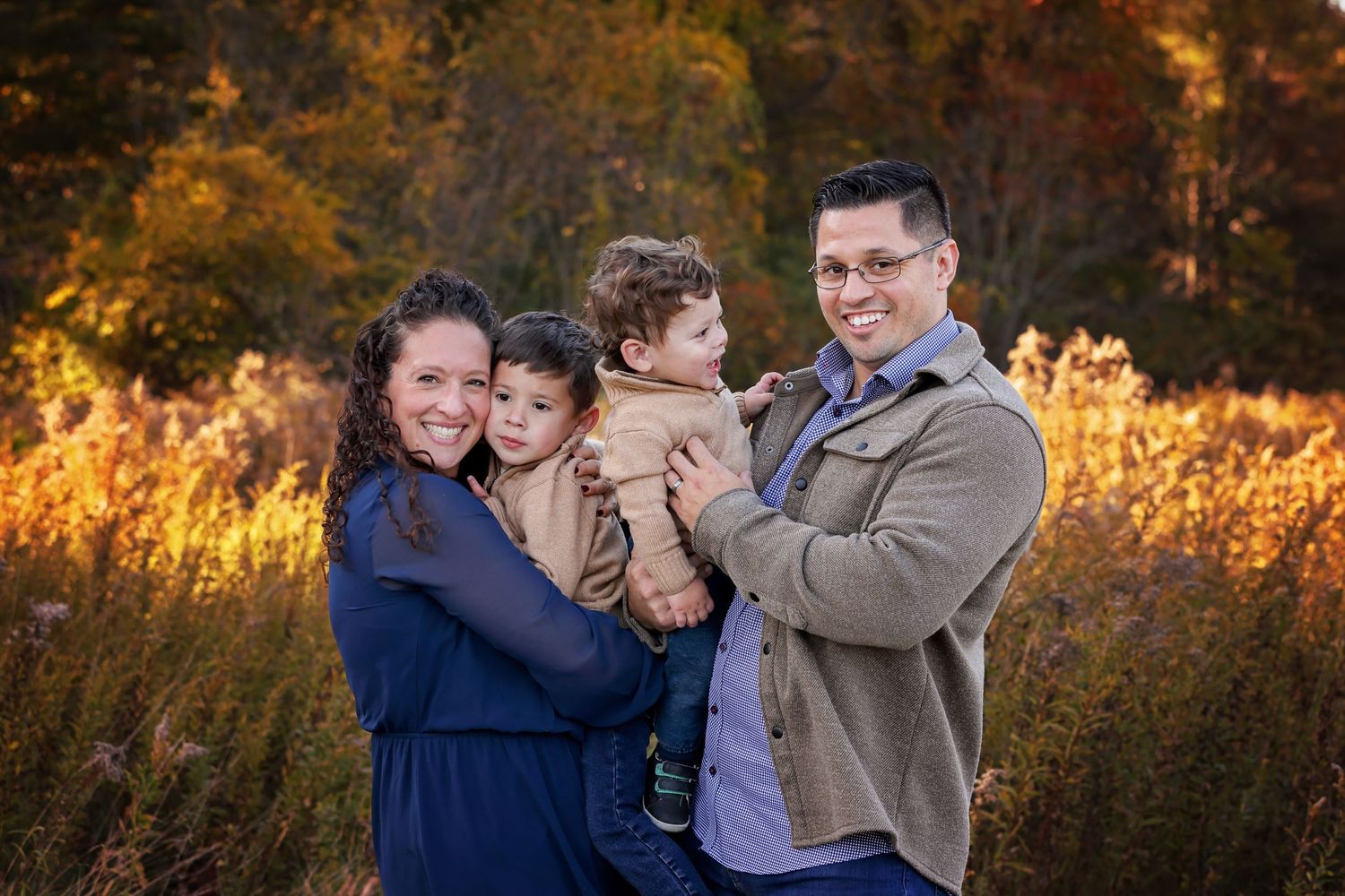 NH Family poses together in a field during golden hour with autumn foliage in the background.